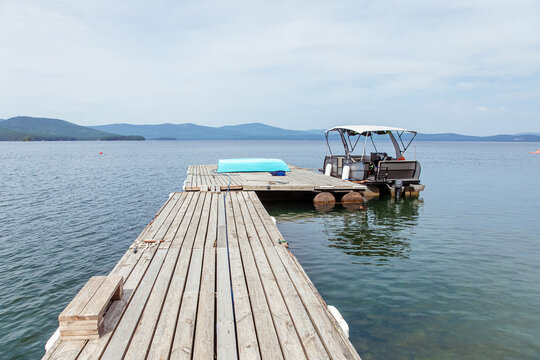 Wooden pier extending into a calm lake with a motorized pontoon boat and a turquoise kayak, surrounded by distant hills and a cloudy sky, perfect for water recreation