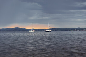 Fototapeta premium Sailboats anchored on Lake under a moody overcast sky, with distant hills and a subtle sunset glow on the horizon, conveying a tranquil yet dramatic atmosphere