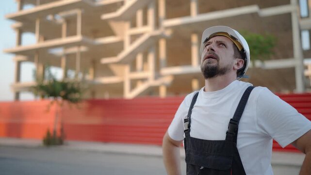 Construction worker wear white safety helmet and work overalls observing progress of residential housing development. Building site inspection, construction industry and project supervision concept.