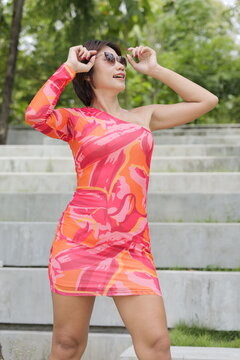An Asian woman in a bright pink and orange patterned one-shoulder dress poses with her hands near her head while standing outdoors on concrete steps