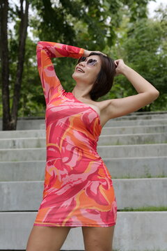 Asian woman wearing sunglasses and a colorful patterned dress posing with hands behind her head in front of concrete park steps