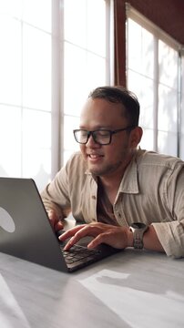 Cheerful young Asian man is sitting down in a cafe while working using his laptop.