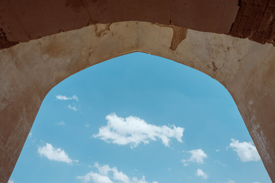Low-angle architectural view from beneath the pointed entrance arch of a historic Safavid caravanserai in Iran, framing a bright blue sky with scattered white clouds.