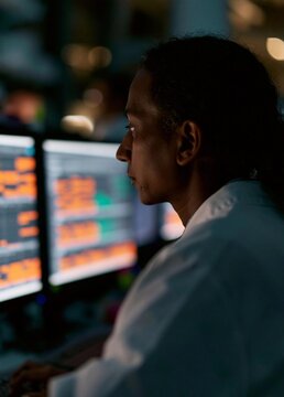 Woman sitting in front of multiple computer monitors in a dark room. she is wearing a white shirt and appears to be focused on the screens.