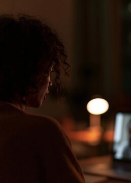 Woman sitting in front of a laptop computer in a dimly lit room. the woman is facing away from the camera, with her back to the viewer. she has curly hair and appears to be deep in thought.