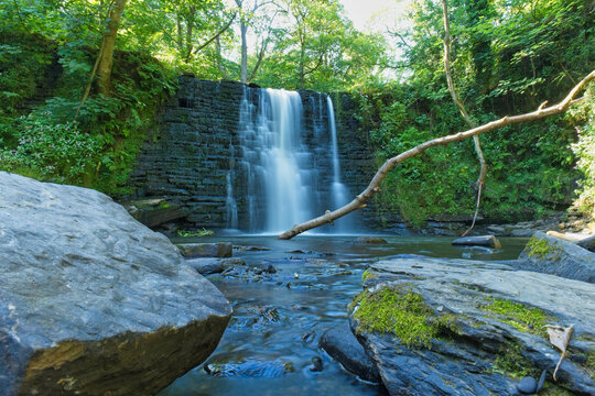A scenic waterfall flows over dark tiered rocks amidst a lush green forest. Large mossy rocks frame a clear stream in the foreground - Ashworth Valley, Rochdale, UK