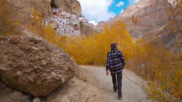 Defocused Indian male tourist wearing hat walking on a small pathway besides the orange colored trees during the autumn season in front of Phugtal monastery in Zanskar Valley, Ladakh, India.