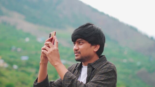 Portrait shot of an Indian male travel influencer shooting videos using his mobile phone for his social media while standing in front of the Himalayan mountains at Manali in Himachal Pradesh, India.