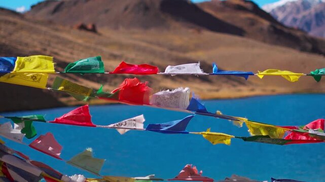 Slow motion shot of colorful Buddhist Tibetan prayer flags waving in the wind in front of blue colored Chandratal lake during the autumn season as seen on the way to Spiti Valley in Himachal, India.
