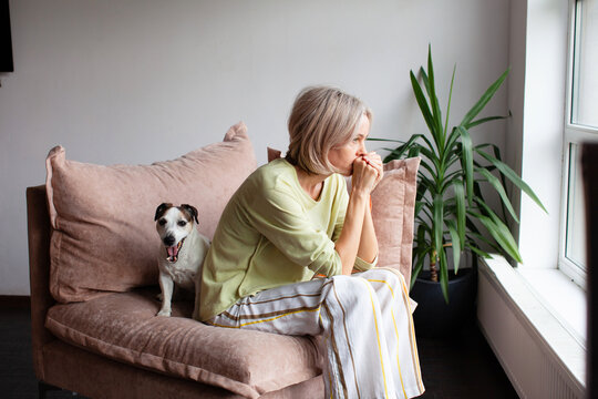 Caucasian woman sitting on couch looking out window with dog. Sad girl feeling lonely at home. Concept of depression or melancholy and support from loyal pet. Emotional domestic scene.