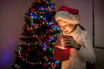 Obraz premium Child opening glowing gift box near decorated Christmas tree. Happy girl wearing Santa hat receiving present. Magic of holiday season and winter celebration at home with light reflection.