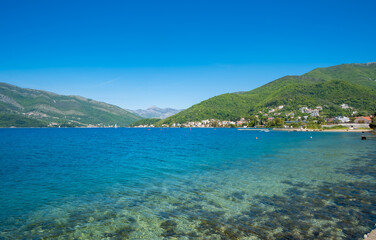 Naklejka premium Clear blue sea meets green hillside under bright blue sky in Tivat, Montenegro, with shoreline featuring small houses and rocky beach.