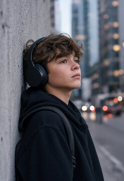 Thoughtful teenage boy in black hoodie listening to music with headphones on city street