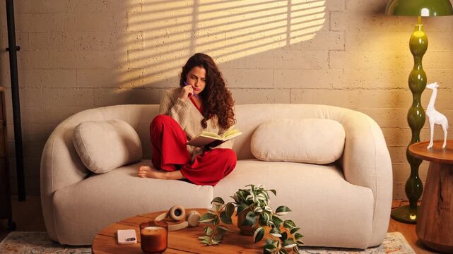 Young woman sitting cross-legged on a cozy couch, holding a notebook and looking away thoughtfully with a pen on her chin. Concept for journaling, creative writing, and self-reflection.
