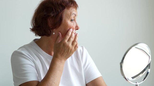 Portrait of elderly woman looking at herself in mirror while sitting at table in bright room. Adult woman examines wrinkles and touches face in cozy room. Concept of care, and enjoyment.