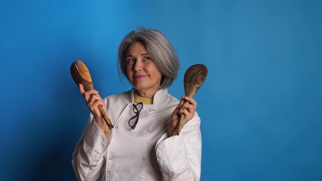 Playful female chef in white uniform holding wooden spoons, dancing and posing fun expressions on a blue background. Concept for passionate cooking, fun recipes, and culinary joy.