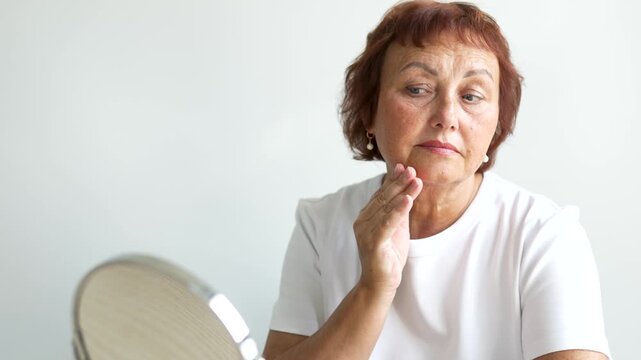 Portrait of elderly woman looking at herself in mirror while sitting at table in bright room. Adult woman examines wrinkles and touches face in cozy room. Concept of care, and enjoyment.