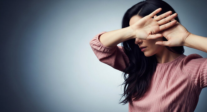 Woman shielding face with hands, dark hair, pink textured garment, soft blue background, concept of shyness or defense