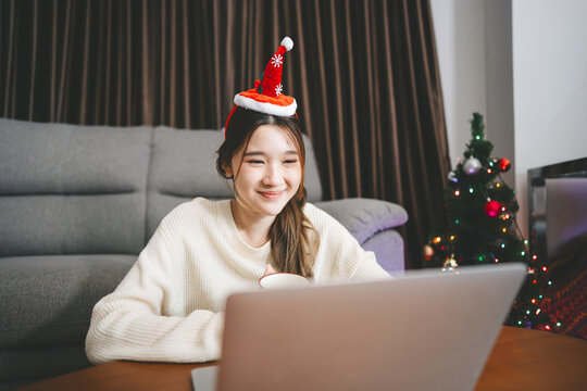 Young adult asian woman using laptop relax sitting in cozy winter living room with christmas tree enjoy alone at home for the holidays