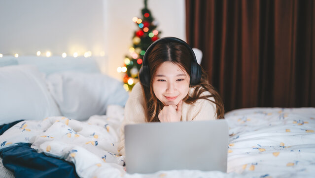 Young adult asian woman using laptop relax sitting in cozy winter bedroom with christmas tree enjoy alone at home for the holidays