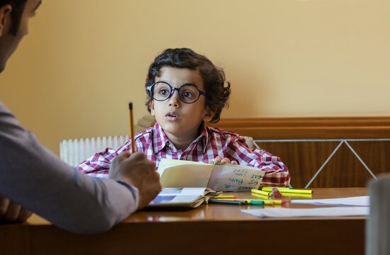 Boy with big round glasses looking at his father while doing homework