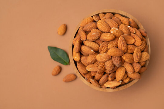 Dried almond kernels in wooden bowl, green leaves and scattered nuts on warm brown background. Almond imagery supports healthy food marketing, nutrition education, packaging and lifestyle content.
