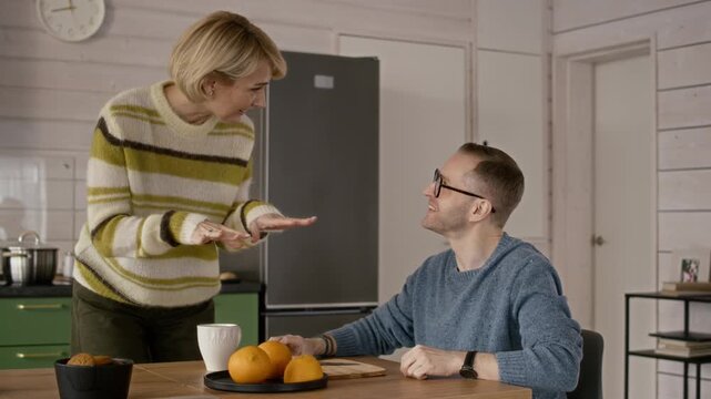 Middle aged Caucasian couple interacting in kitchen, woman showing smartphone screen to man drinking coffee. Ideal for mobile app marketing or e-commerce campaign
