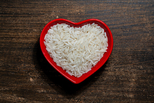 Top view of raw white rice grains inside red heart-shaped bowl on rustic wooden table, closeup. Ideal for concepts about healthy eating, love for cooking, charity, and agricultural trade