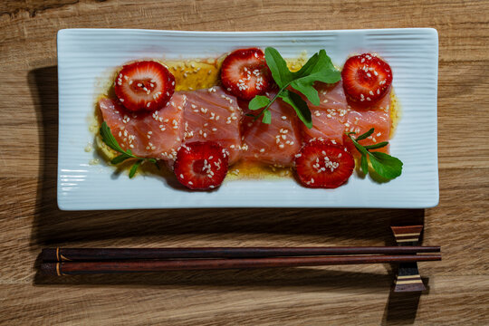 Top view of fresh salmon slices served with strawberry, sesame seeds, and arugula on white plate, closeup. Perfect for gourmet restaurant menus, fusion cuisine, and luxury food marketing