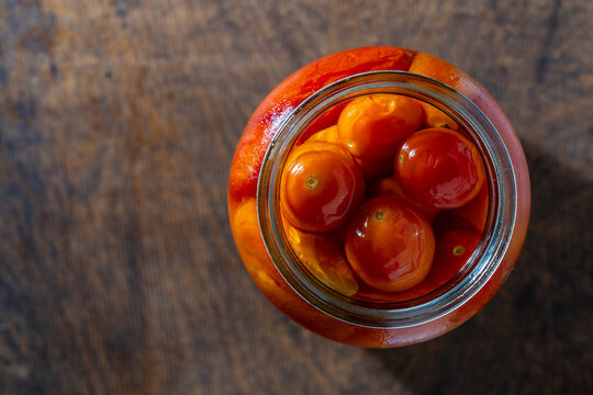 Top view of an open glass jar filled with marinated red cherry tomatoes on rustic wooden table, closeup. Perfect for autumn harvest, preservation recipes, and organic homemade food concepts