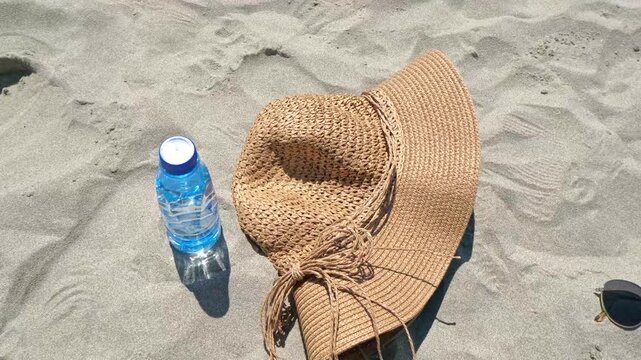 A blue water bottle and a large straw hat resting on sandy beach. The scene suggests relaxation and summer activities.