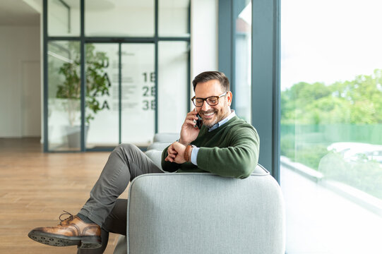 Business professional communicating using a mobile phone, smiling while checking a wristwatch, sitting comfortably in an office reception area next to a window