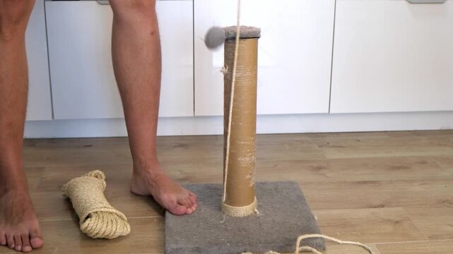 A young Caucasian man repairs a cat scratching post. He stands barefoot next to the post, with tools and materials scattered on the floor.