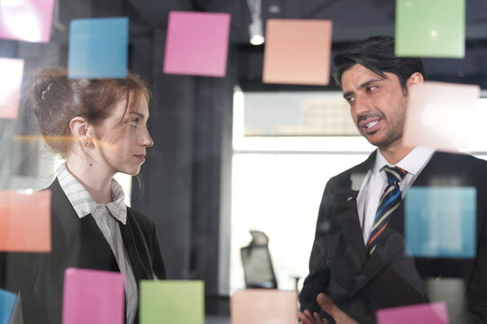 Manager makes a skeptical face at a sticky note while the young woman in profile keeps scanning the colorful plan on the glass wall in a bright modern coworking office.