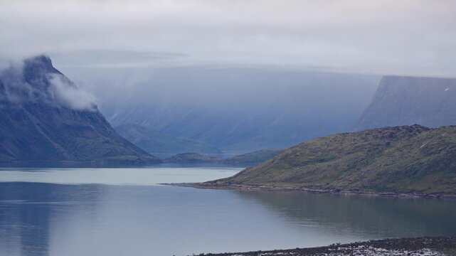 Atmospheric landscape view of dramatic, fog-shrouded mountain summits rising above calm fjord waters leading toward Auyuittuq National Park.