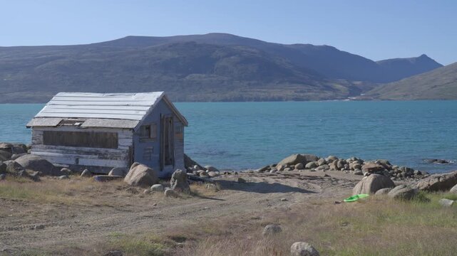 Wide ground shot capturing a small, weathered wooden outpost cabin situated along the rocky shoreline overlooking sunny turquoise fjord waters.