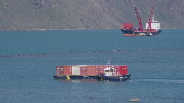 Wide telephoto shot tracking a tugboat maneuvering a red container barge across deep blue fjord waters with a large crane-equipped sealift freighter anchored in the upper frame.