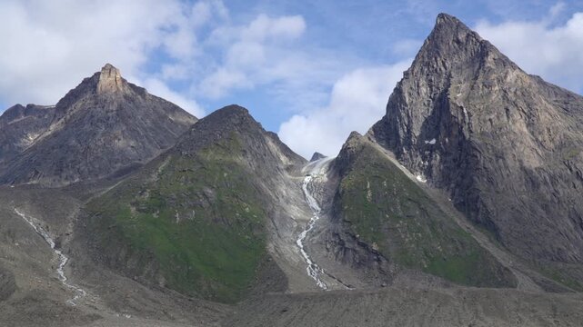 High-angle shot revealing the toe of a receding glacier tucked within a rugged mountain valley, highlighting glacial retreat, exposed bedrock, and moraine sediment in Auyuittuq National Park.