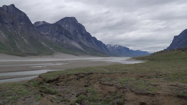 Wide horizontal shot along the banks of a glacial river bed framed by steep mountain cliffs in the Akshayuk Pass corridor.