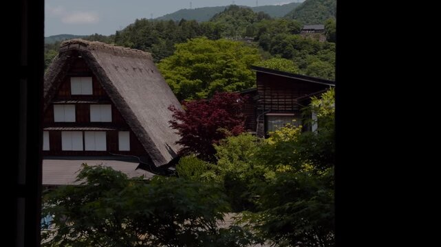 View through a dark wooden window frame of a traditional gassho-zukuri farmhouse with a steep thatched roof, surrounded by lush green trees and forested mountains.
