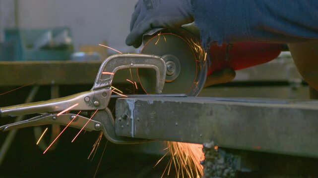 Angle grinder sparks workbench clamp tool cutting metal, handheld close up shot. Bright fiery shower illuminating steel piece secured on workshop table.