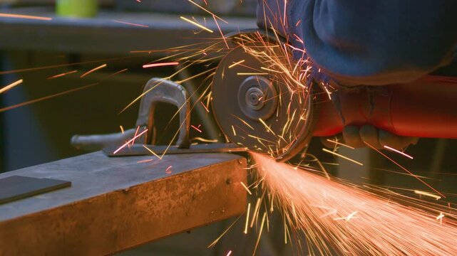 Angle grinder sparks blacksmith gloved hand cutting metal, handheld close up shot. Heavy machinery tool working on steel workshop component making bright fire.