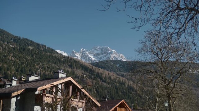 Snowy mountain peak houses roofs forest landscape, handheld shot. Winter alpine range with white summit and dry tree ridges behind village chalets.