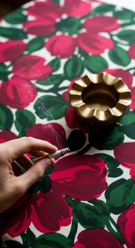 Hand holding a burning cigarette over a floral patterned surface with an ashtray, symbolizing habit, pause, or reflection