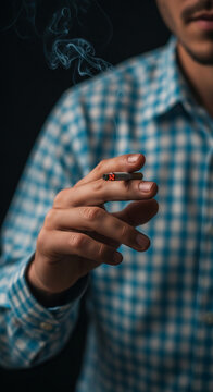 A man's hand holds a lit cigarette with smoke rising, set against a dark background and a checkered shirt, symbolizing habit or contemplation.