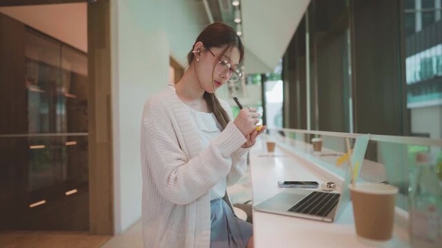 Young adult business asian woman using laptop wear eyeglasses using pen writing sticky note