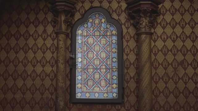 A small diamond patterned stained glass window glows softly in a dim, richly decorated chapel interior in Paris.