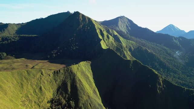 Volcanic mountain ridge with jagged sunlit peaks and a wide grassland plateau overlooking distant hazy summit tropical highland terrain aerial drone footage