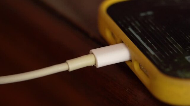 Unrecognizable woman lightning cable into smartphone connector to charge sitting at home table.