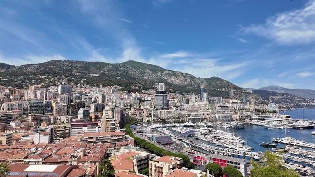 Panoramic view of Monaco and Port Hercule with luxury yachts, modern skyline, and Mediterranean coastline framed by dramatic mountains under a bright blue sky on a sunny day.